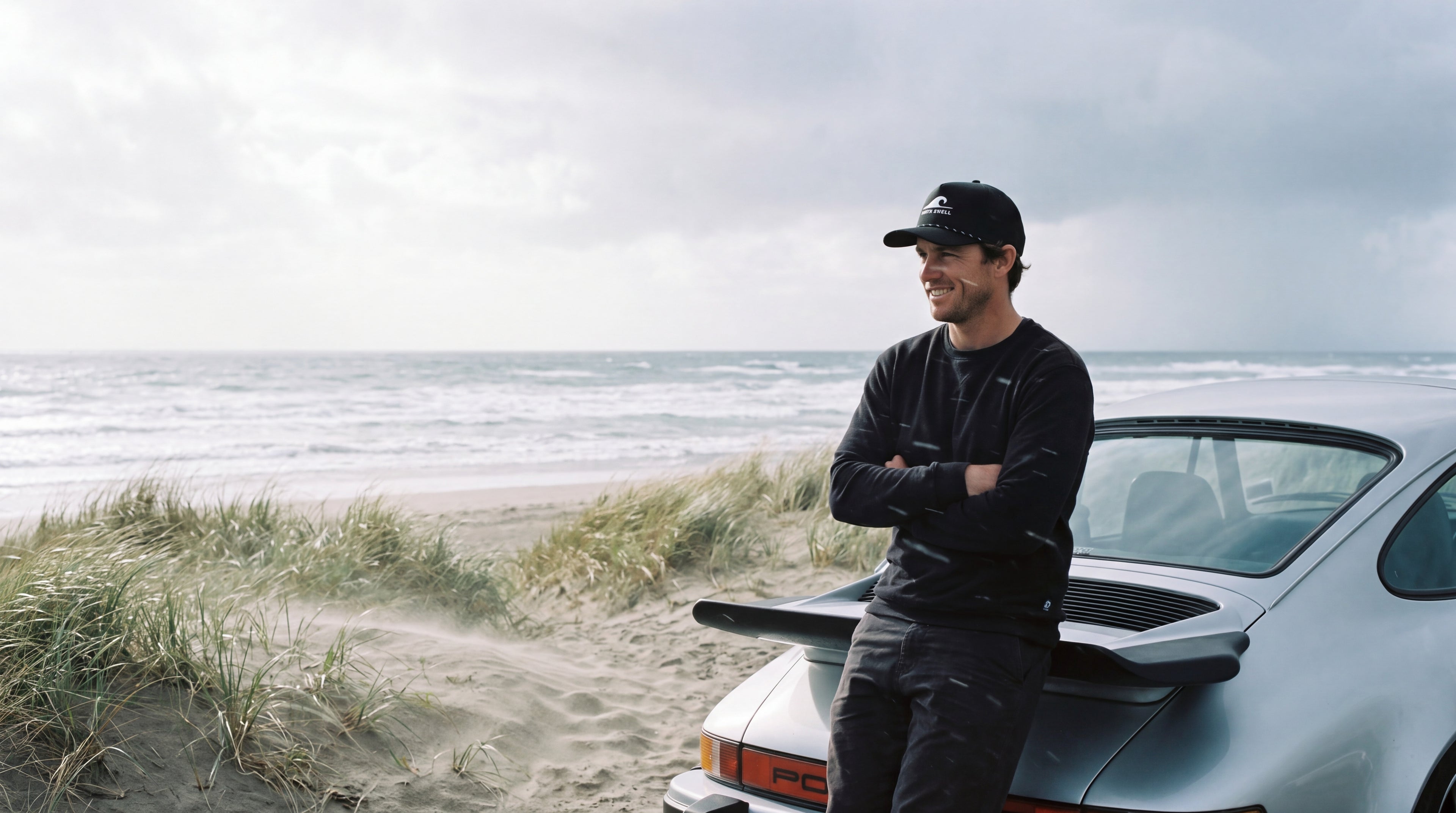 A man in a black North Swell Swell Seeker snapback leaning against a classic sports car at a Northland beach car park